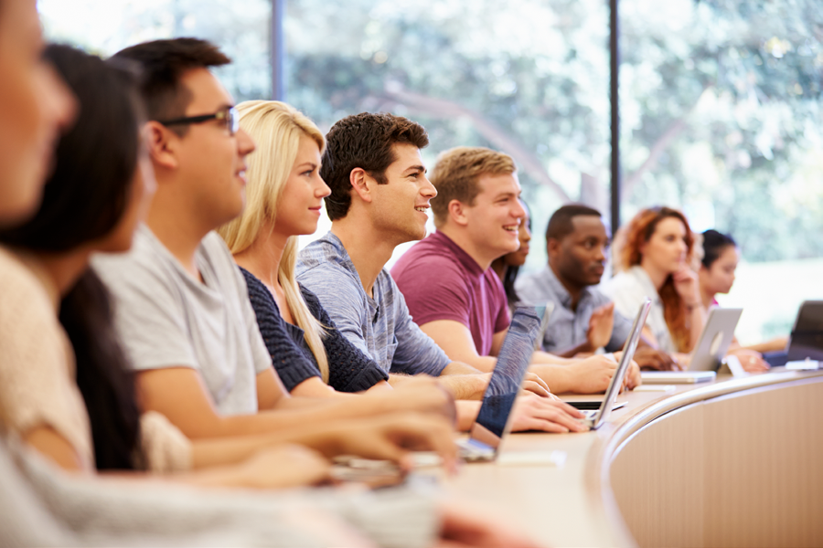 A class of smiling college students inside the classroom listening to a lecture.